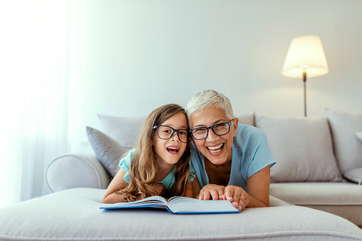 Loving grandmother teaching granddaughter holding book laying on sofa, grandma baby sitter embracing kid girl reading fairytale to cute child, nanny granny telling story to preschool grandchild while laying on tummy on sofa during the day.