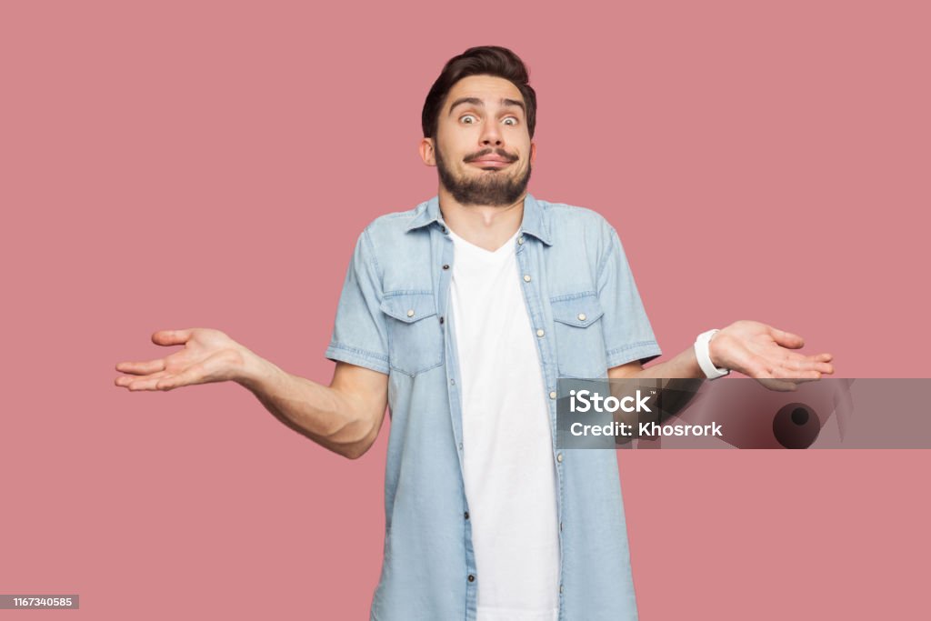I don't know. Portrait of confused handsome bearded young man in blue casual style shirt standing with raised arms and looking at camera with answer. I don't know. Portrait of confused handsome bearded young man in blue casual style shirt standing with raised arms and looking at camera with answer. indoor studio shot, isolated on pink background. Men Stock Photo