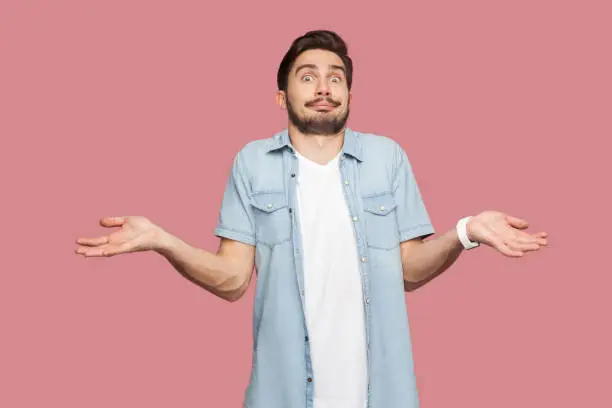 I don't know. Portrait of confused handsome bearded young man in blue casual style shirt standing with raised arms and looking at camera with answer. indoor studio shot, isolated on pink background.