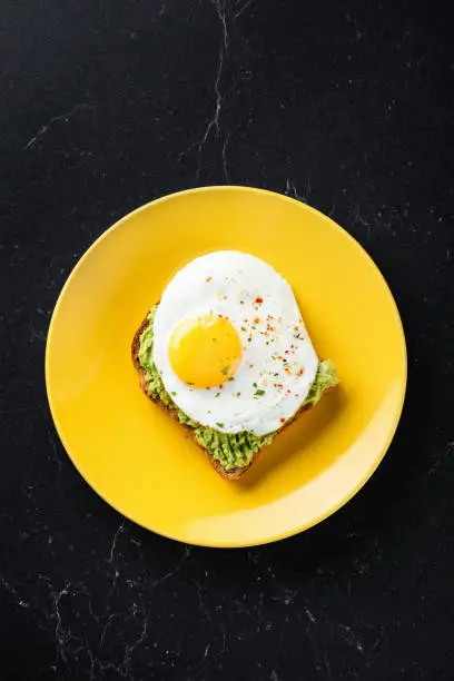Sunny side up egg and mashed avocado on toasted sandwich bread. Healthy breakfast lunch or snack. On yellow plate, black marble table background, top view