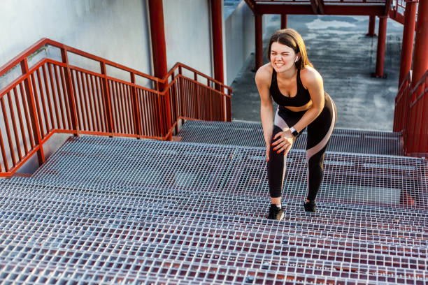 portrait of young sporty girl in black sportwear standing on stairs and have strong hurt problem with knee, spasm painful. gripping leg with hand with clenching teeth. - running athlete staircase teenager imagens e fotografias de stock