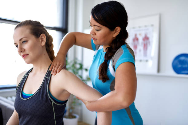 Chinese woman physiotherapy professional giving a treatment to an attractive blond client in a bright medical office Candid image of an attractive patient during a appointment with professional asian physiotherapist in design kinesio clinic working and massaging her neck and shoulder pain sports physical stock pictures, royalty-free photos & images