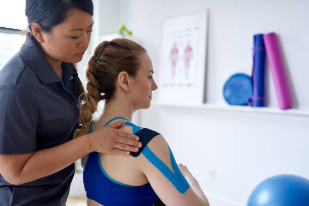 Chinese woman massage therapist applying kinesio tape to the shoulders and neck of an attractive blond client in a bright medical office Candid image of an attractive patient during appointment with a professional asian physiotherapist in design kinesiotherapy clinic stretching medical kinesio tape for neck and shoulder pain. sports medicine stock pictures, royalty-free photos & images