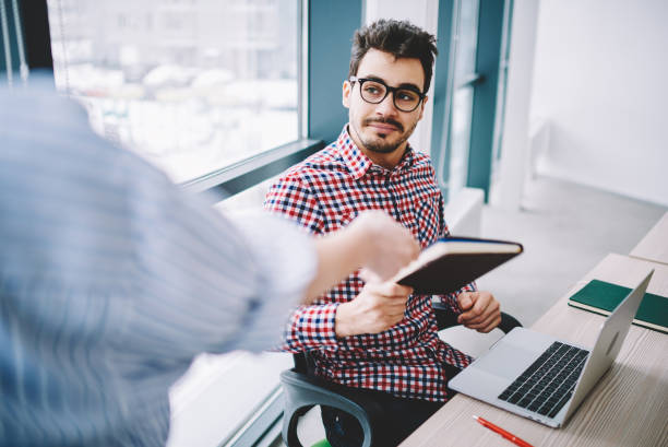 istock Handsome man office worker in trendy eyeglasses for vision correction sitting at desktop with new netbook and taking notebook from colleague, cropped back view of male with textbook with information 1167366761