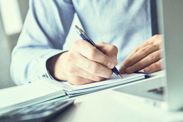 istock Businessman hand writing note on a notebook. Business man working at office desk. Close up of empty notebook on a blackboard with office supplies. 1167376605