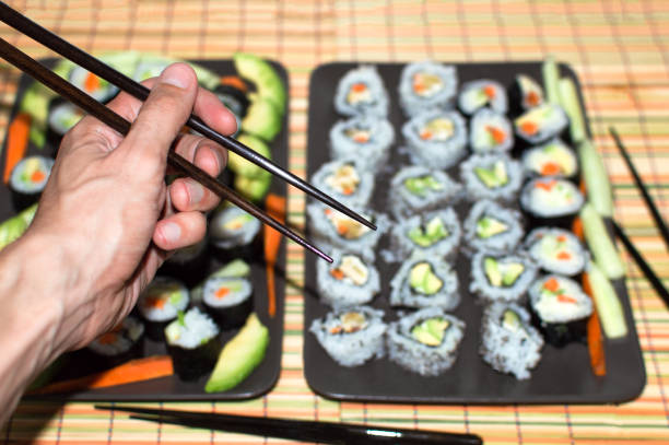 Hand using chopsticks with background full of different types of sushi to eat. Choosing and eating traditional sushi with japanese decoration.