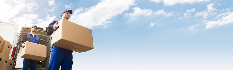 Close-up Of Two Young Delivery Men Carrying Cardboard Box In Front Of Truck