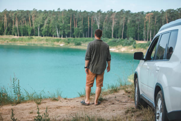 человек возле белого автомобиля suv на краю глядя на озеро с голубой водой - waters edge lake beach tree стоковые фото и изображения