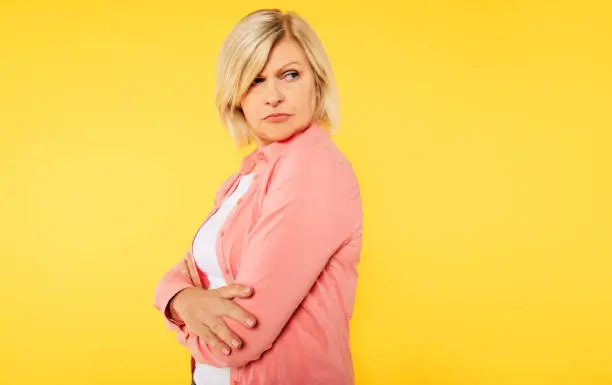 Photo of Angry blonde senior woman with crossed arms is posing over yellow background