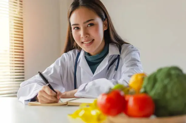 Photo of young nutritionist female doctor or medical looking at camera writing diet plan and fresh vegetables, fruits on desk at laboratory office, nutrition, healthy lifestyle, healthy food, dieting concept