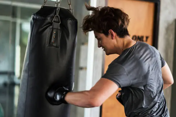 American young boxer punching hard at big bag at fitness gym. Athletic man training Thai boxing punch for bodybuilding and healthy lifestyle concept. Energy guy workout in sport club.