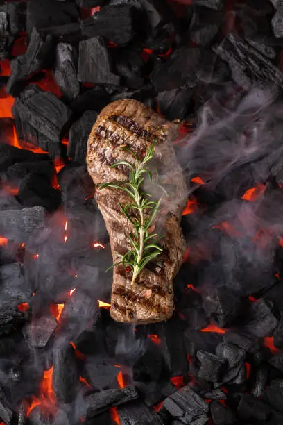 Fried meat steak on a black background of charcoal. Cooked juicy steak with smoke on the coals with rosemary. Grilled delicious marinated steak on coals, closeup photo. Top view