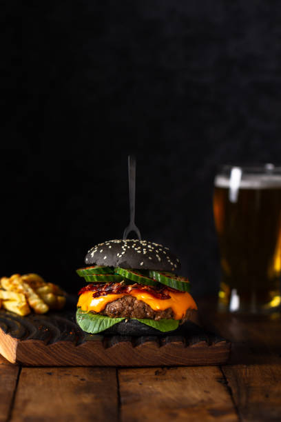 black burger with beef served on wooden chopping board over wooden table with black background. black burger with french fries and beer. - salad food and drink food lettuce imagens e fotografias de stock