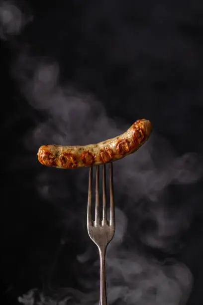 Grilled sausage on a fork fuming smoke dark black background. German sausage. Nuremberg sausage on fork