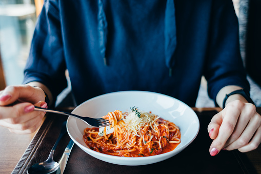 Close-up of young woman eating Italian pasta at table in restaurant holding fork. Plate of delicious spaghetti with tomato sauce and cheese.