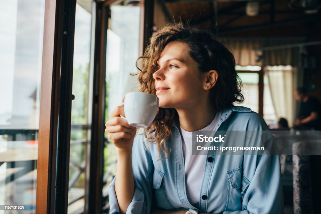 Smiling calm young woman drinking coffee Smiling calm young woman drinking coffee looking out the window sitting at table in restaurant. Coffee - Drink Stock Photo
