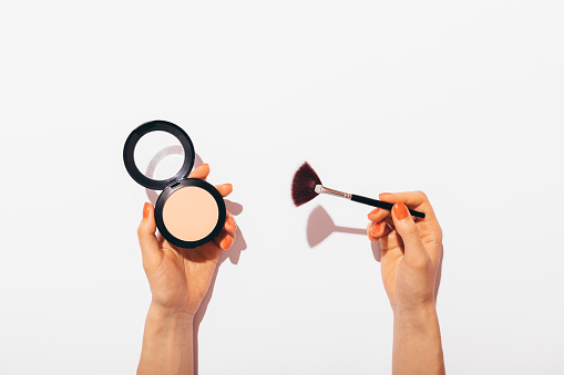 Female hands holding pressed powder and makeup brush on white background, flat lay concept composition with hard light.