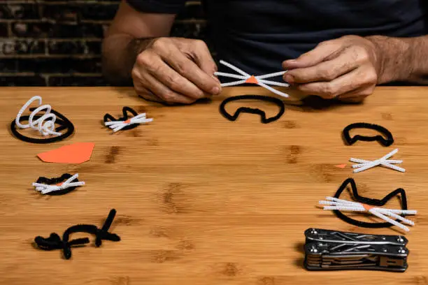 This image features not only the crafts, but plenty of negative space for copywriting. Halloween pipe-cleaner and construction paper crafts featuring Black Cats.  A man's hands in the frame show the progress of the crafts project a bit.