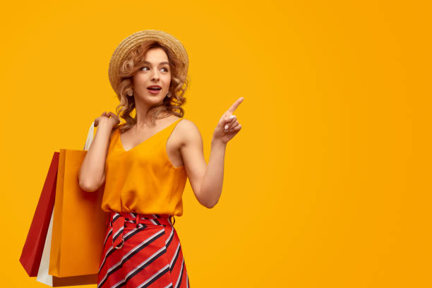 Stylish shopaholic with purchases pointing at empty space Surprised trendy young woman with paper bags pointing at blank space and looking away after shopping against vivid yellow background young women shopping stock pictures, royalty-free photos & images