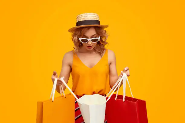 Shocked young woman in trendy outfit looking at purchases inside paper bag during shopping against vivid yellow background