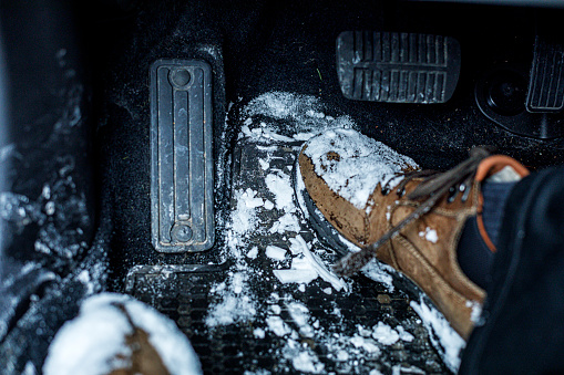 Personal perspective high angle view looking down at the driver's frozen snow covered winter boots which are leaving a sloppy mess on the flooring carpet of a car near the brake pedal and gasoline accelerator pedal. Part of a 