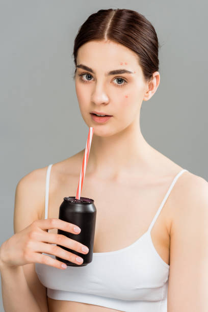 young woman with acne on face holding can with straw isolated on grey