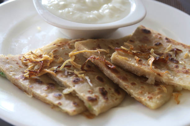 Stock photo of North Indian breakfast dish, slices of aloo parathas with sprinkle of caramelised onion on top served in a white plate with thick yogurt / curd traditional Indian recipe. Savoury bread stuffed with garam masala potatoes and onions. imagen del buffet de desayuno del norte de la india, rebanadas de aloo parathas con espolvorear de cebolla caramelizada en la parte superior en plato blanco, pan salado relleno de patatas garam masala, restaurante del hotel casero aloo paratha plato, raita
