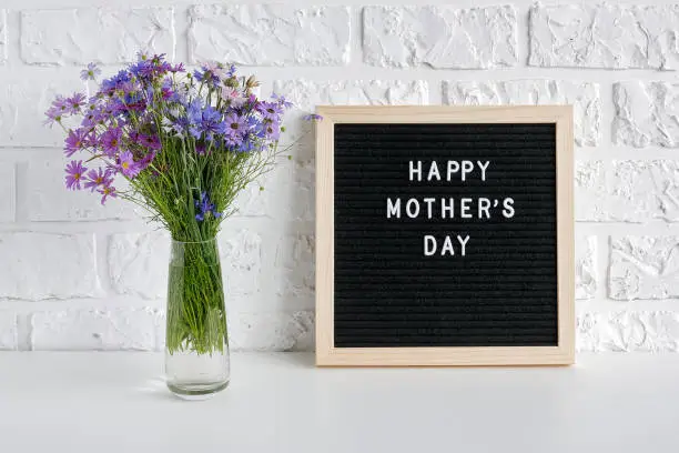 Photo of Happy mothers day text on black letter board and bouquet blue flowers in vase on table against white brick wall. Concept Mothers Day. Template for greeting card, postcard