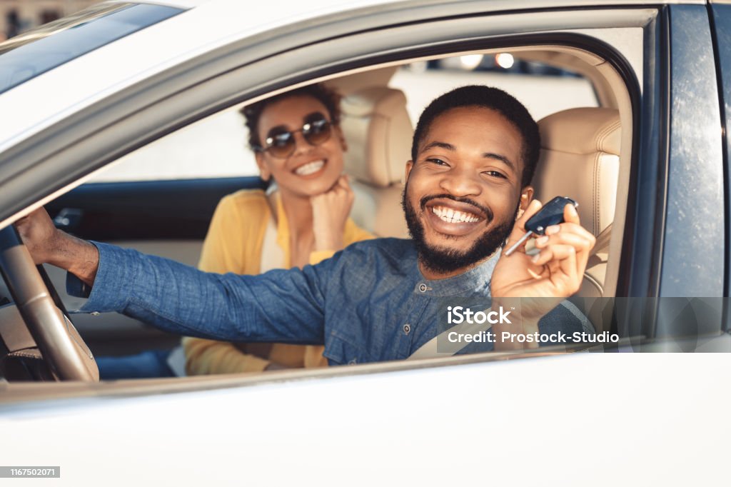 Visiting car dealership. Afro couple showing car key Visiting car dealership. Afro couple showing car key, looking at camera and smiling, sitting in new car Car Stock Photo