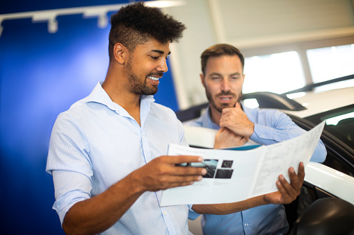 Car salesperson holding a brochure and informing an interested customer in a dealership showroom.