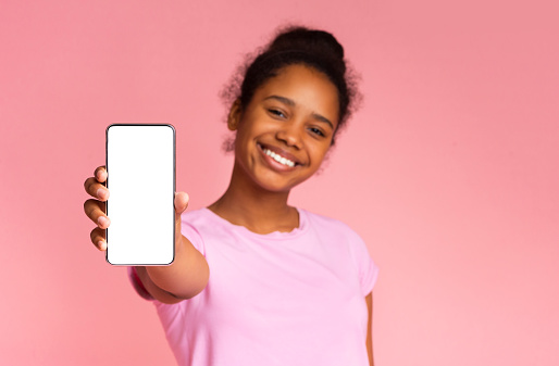 Cute african american girl showing blank smartphone screen, perfect for educational application or website. Pink background with empty space.