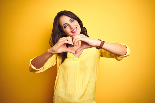 Beautiful elegant woman standing over yellow isolated background smiling in love showing heart symbol and shape with hands. Romantic concept.