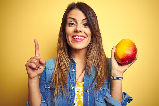 Young beautiful woman eating fresh healthy mango over yellow background surprised with an idea or question pointing finger with happy face, number one