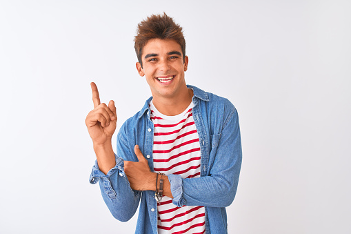 Young handsome man wearing striped t-shirt and denim shirt over isolated white background with a big smile on face, pointing with hand and finger to the side looking at the camera.