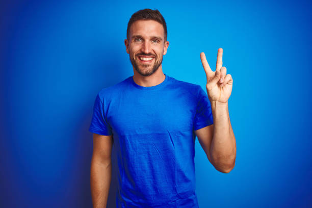 Young handsome man wearing casual t-shirt over blue isolated background smiling with happy face winking at the camera doing victory sign. Number two. Young handsome man wearing casual t-shirt over blue isolated background smiling with happy face winking at the camera doing victory sign. Number two. young man wink stock pictures, royalty-free photos & images