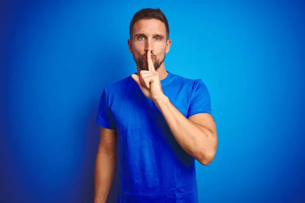 Photo of Young handsome man wearing casual t-shirt over blue isolated background asking to be quiet with finger on lips. Silence and secret concept.