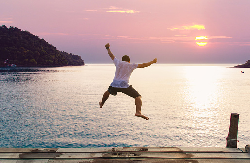 Young man jumping into the sea from wooden jetty on a beach. Happy summer vacation concept.