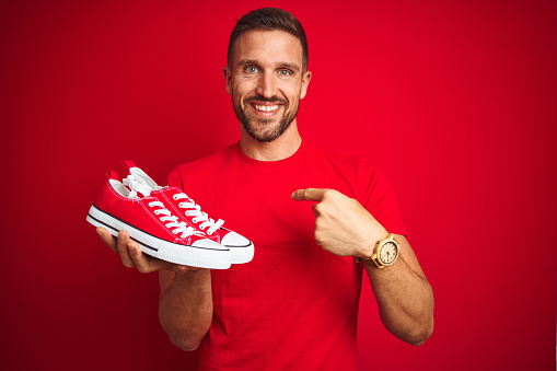 Young man holding casual sneakers shoes over red isolated background with surprise face pointing finger to himself