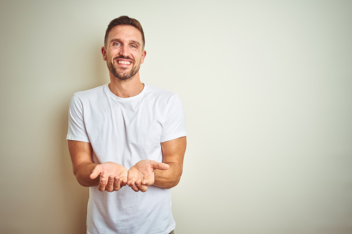 Young handsome man wearing casual white t-shirt over isolated background Smiling with hands palms together receiving or giving gesture. Hold and protection