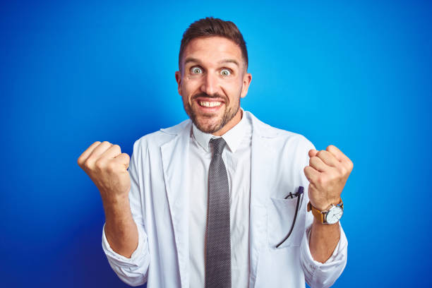 istock Young handsome doctor man wearing white professional coat over isolated background celebrating surprised and amazed for success with arms raised and open eyes. Winner concept. 1167539208