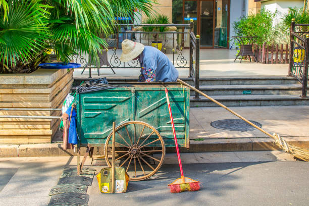 Haikou, China - May 12, 2019: a cleaner with a makeshift trolley cleans the garbage near the hotel. Broom with a bamboo handle. The cart has original wheels. An unexpected contrast of this scene with the stereotype of fast-progressing China.