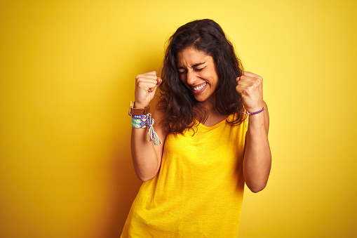 Young beautiful woman wearing t-shirt standing over isolated yellow background very happy and excited doing winner gesture with arms raised, smiling and screaming for success. Celebration concept.