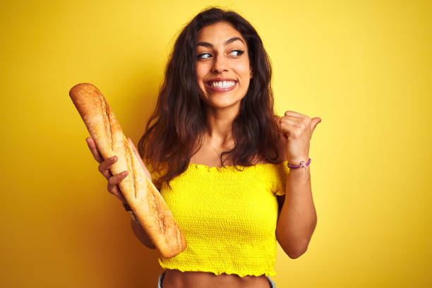 istock Young beautiful baker woman holding bread standing over isolated yellow background pointing and showing with thumb up to the side with happy face smiling 1167548557