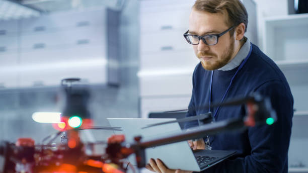 istock Male Engineer Programs Drone while Holding Laptop in His Hands. He Works in a Bright Modern High-Tech Laboratory. 1167549759