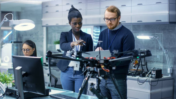 coordenadores fêmeas masculinos e pretos caucasianos que trabalham em um projeto do zangão com ajuda do portátil e de tomar notas. ele trabalha em um laboratório de alta tecnologia moderna e brilhante. - engineer occupation women industrial - fotografias e filmes do acervo