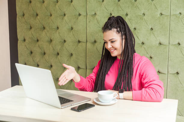 retrato de joven feliz con dreadlocks negros peinado en blusa rosa sentada y hablando con la cámara web en el ordenador portátil y el saludo con la sonrisa dentada, dando la mano a la oferta en línea. - turkish ethnicity white black contemporary fotografías e imágenes de stock