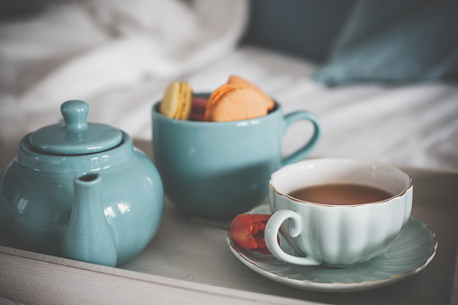 White teapot, mug of herbal beverage in cozy home interior. Drinks, book with flower are on wooden serving tray. Breakfast on sofa with lace cover. Still life for tea time.