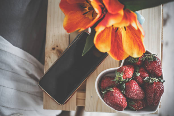 Copy space shot of delicious snack set up on a bedside table. Heart shaped bowl filled with fresh strawberries lying next to a mobile phone and a vase with beautiful tulips that are blooming.