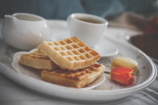 Close up shot of a plate with sweet waffles and coffee served on a tray as a breakfast in bed. There is also a single macaroon and a tulip for decoration.