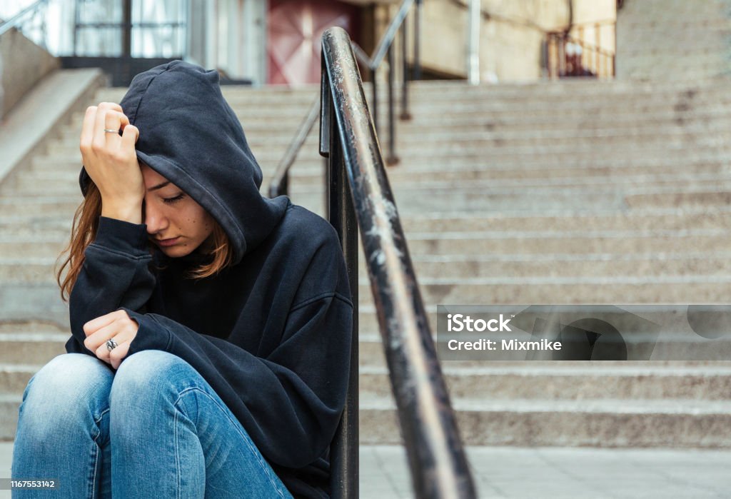 Depressed young woman sitting on the street Teenage girl sitting outdoors on the staircase covering her head with sweatshirt hood Teenager Stock Photo